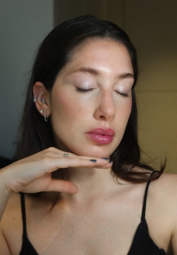 Femme aux yeux fermés et aux lèvres roses brillantes posant avec la main sous le menton, portant un haut spaghetti noir et plusieurs piercings aux oreilles.