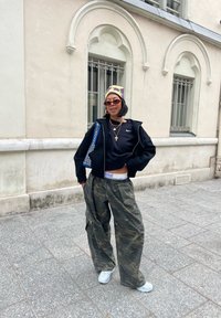 Sweat à capuche noir court, pantalon cargo camouflage, et baskets blanches. Accessoires comprenant des lunettes de soleil et des colliers en or superposés. Sac à motifs bleus.