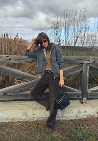 Szara bluza z zamkiem i kieszeniami z przodu, musztardowy t-shirt, ciemnobrązowe spodnie ze sztruksu oraz czarna torebka na tle naturalnego krajobrazu.