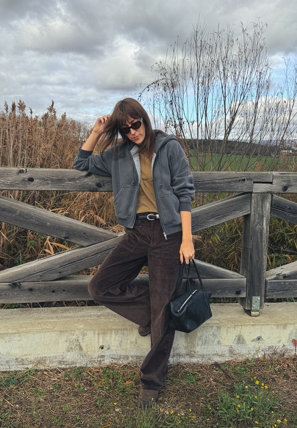 Hanorac gri cu fermoar și buzunare frontale, tricou muștar, pantaloni din catifea maro închis și geantă neagră, pe un fundal natural.