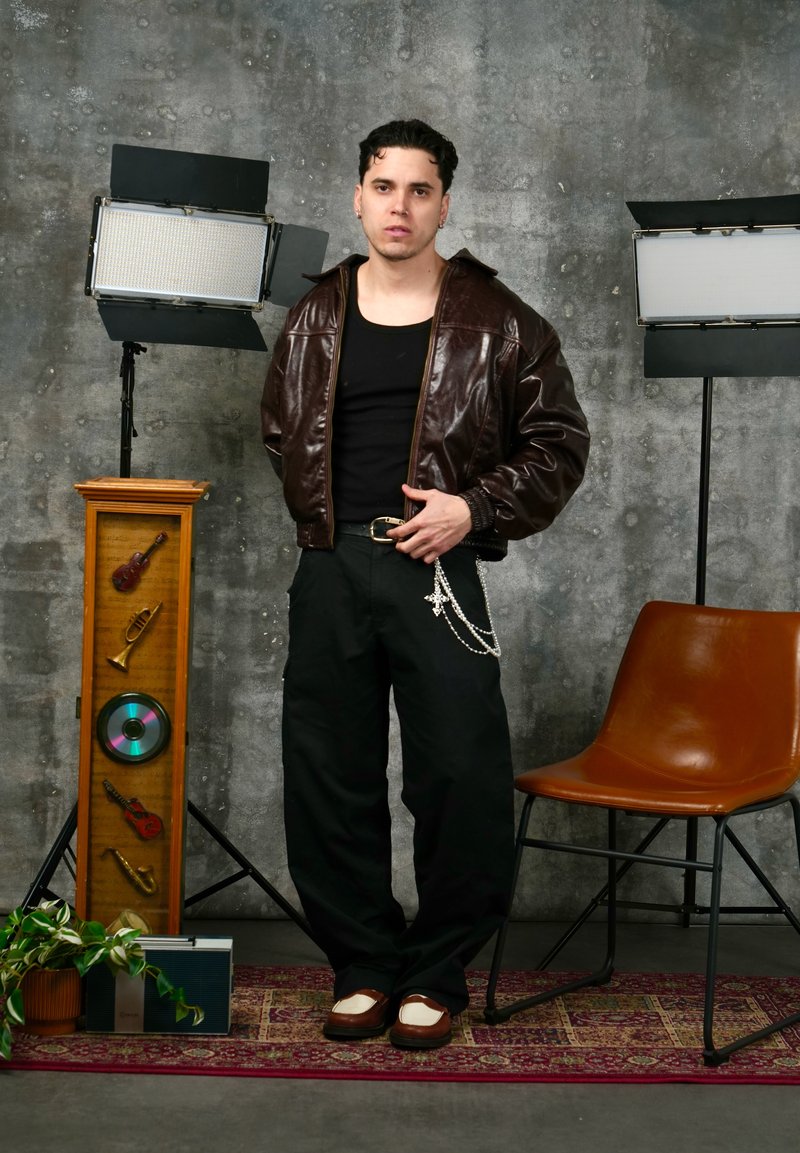 Un tânăr cu o geacă de piele maro și pantaloni negri stă pe un covor cu model, între două lumini de studio și un fotoliu maro.