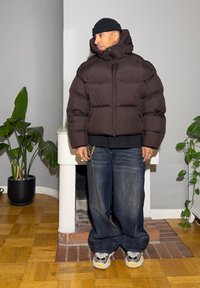 Ruskea toppatakki, jossa korkea kaulus, yhdistetty löysiin sinisiin farkkuihin ja monivärisiin lenkkareihin. Sisätiloissa, joissa on ruukkukasveja.