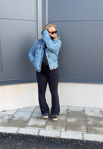 Chaqueta de mezclilla sobre una blusa negra, combinada con pantalones de campana negros. Zapatillas negras con suelas de color claro. Fondo gris con pavimento.