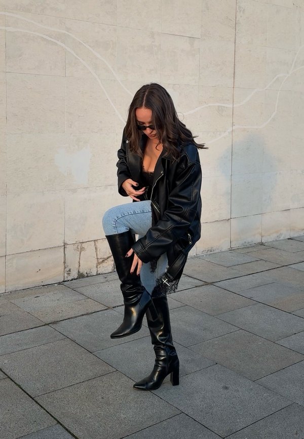 Nő napszemüvegben, fekete bőrdzsekiben és fekete magas sarkú csizmában, a jobb csizmáját igazítja, miközben csempézett járdán áll.