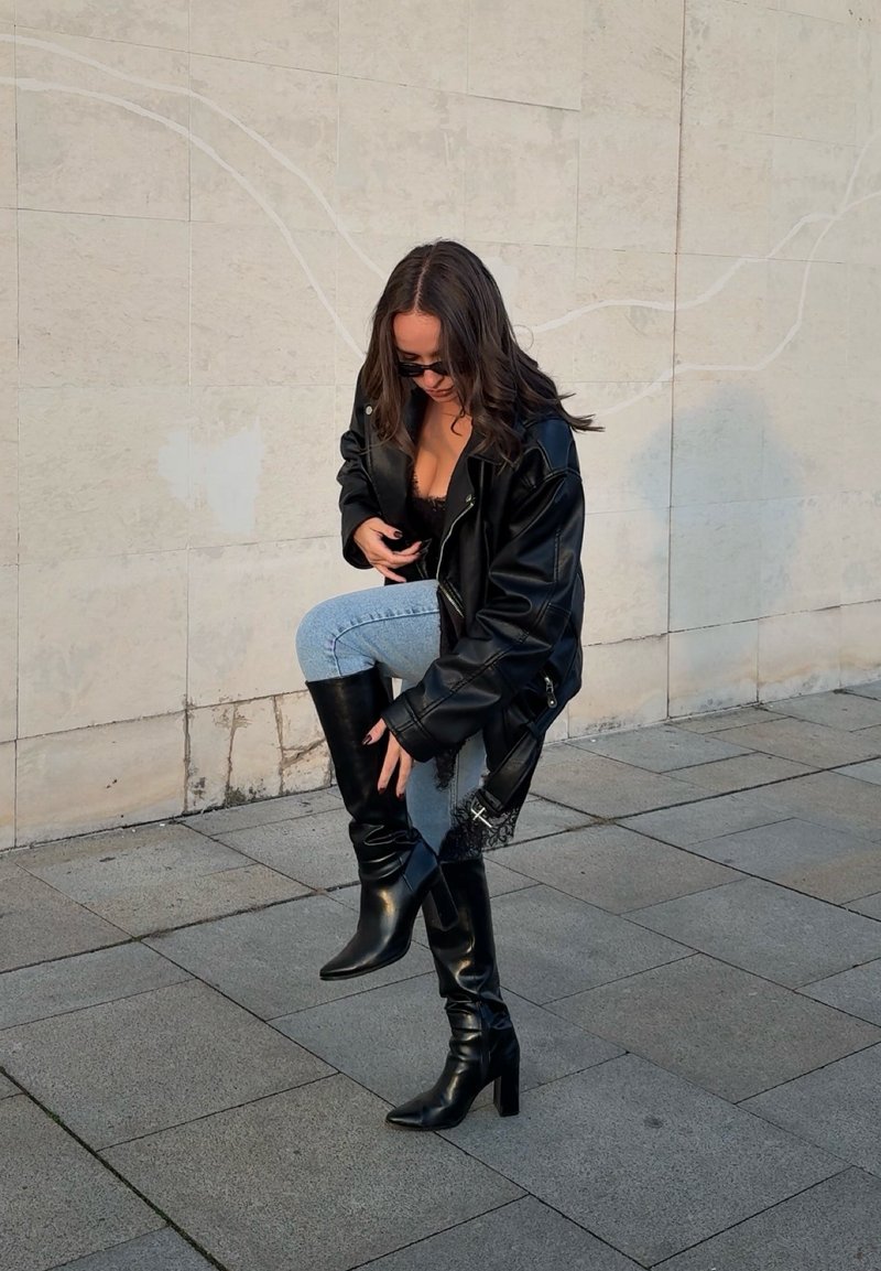 Donna con occhiali da sole, giacca di pelle nera e stivali neri con tacco, mentre aggiusta lo stivale destro in piedi su un pavimento piastrellato.