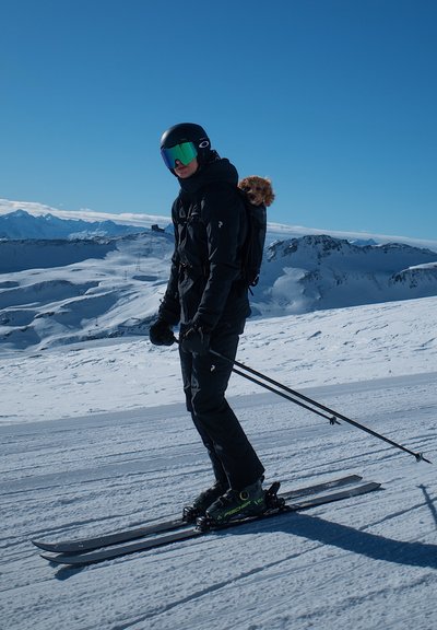 Equipo de esquí en una pendiente cubierta de nieve: chaqueta y pantalones negros, botas de esquí con acentos verdes, bastones de esquí y un perro pequeño en una mochila. Cielo azul brillante.