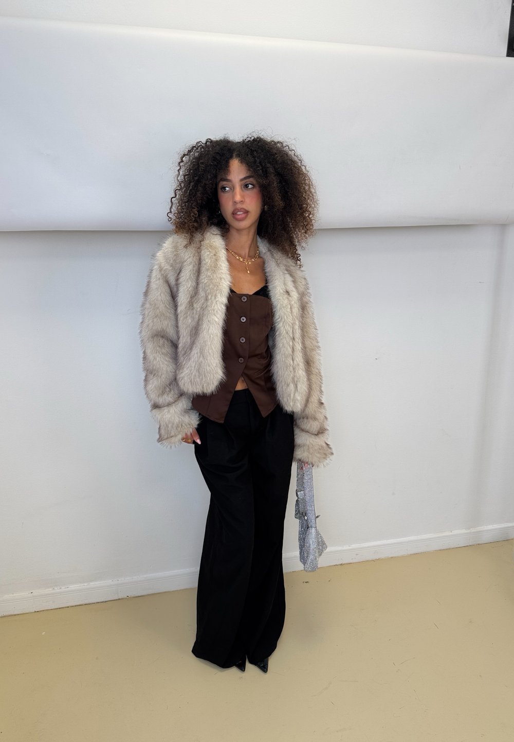 Chaqueta de pelo sintético gris esponjoso sobre una blusa marrón de botones en el frente, combinada con pantalones negros holgados. Sujetando un bolso texturizado. Fondo neutro.