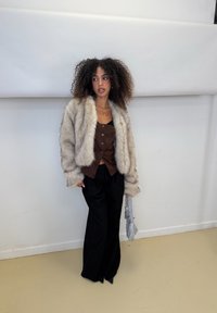 Chaqueta de pelo sintético gris esponjoso sobre una blusa marrón de botones en el frente, combinada con pantalones negros holgados. Sujetando un bolso texturizado. Fondo neutro.
