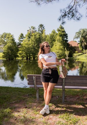 Camiseta blanca con bordados florales, shorts de mezclilla negros, cinturón de leopardo, zapatillas blancas y un bolso beige con un patrón, sentado en un banco.