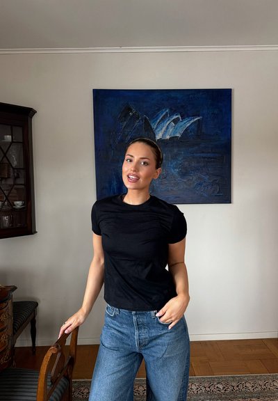 Joven con camiseta negra y jeans azules de pie junto a una silla de madera en una habitación con una pintura abstracta azul oscuro en la pared.