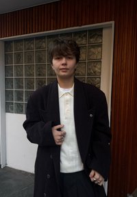 Manteau noir oversize à manches larges porté sur un polo cr ème clair. Col plié avec détail à double bouton. Porte plusieurs bagues en argent.