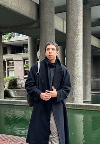 Casaco duplo em preto sobre uma blusa escura, calças cinzentas e acessórios com anéis brilhantes. O fundo apresenta colunas de concreto e água verde.