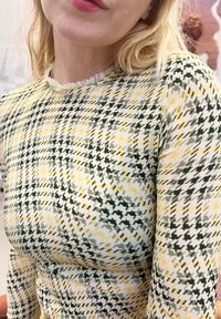 Primo piano di una persona che indossa una maglia con motivo pied-de-poule giallo, verde e bianco, con collo arricciato e maniche lunghe.