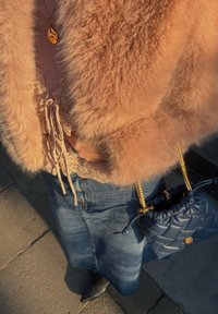 Persoană purtând o jachetă roz pufoasă, top cu dantelă, fustă din denim, ținând o geantă albastră matlasată cu bretele din lanț auriu.