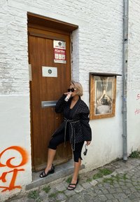 Svart mønstrete oversized jakke med innsnørt midje, kombinert med svarte cropped bukser og svarte slidesandaler, stående ved en tre dør.