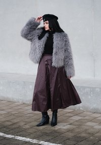 Veste en fausse fourrure grise, haut côtelé noir, jupe midi en satin bordeaux, et bottines noires. Modèle portant un béret noir, debout sur le trottoir.