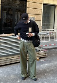 Hombre con pantalones verdes holgados y sudadera negra sosteniendo una taza de café, de pie en una calle con tubos de construcción de metal detrás de él.