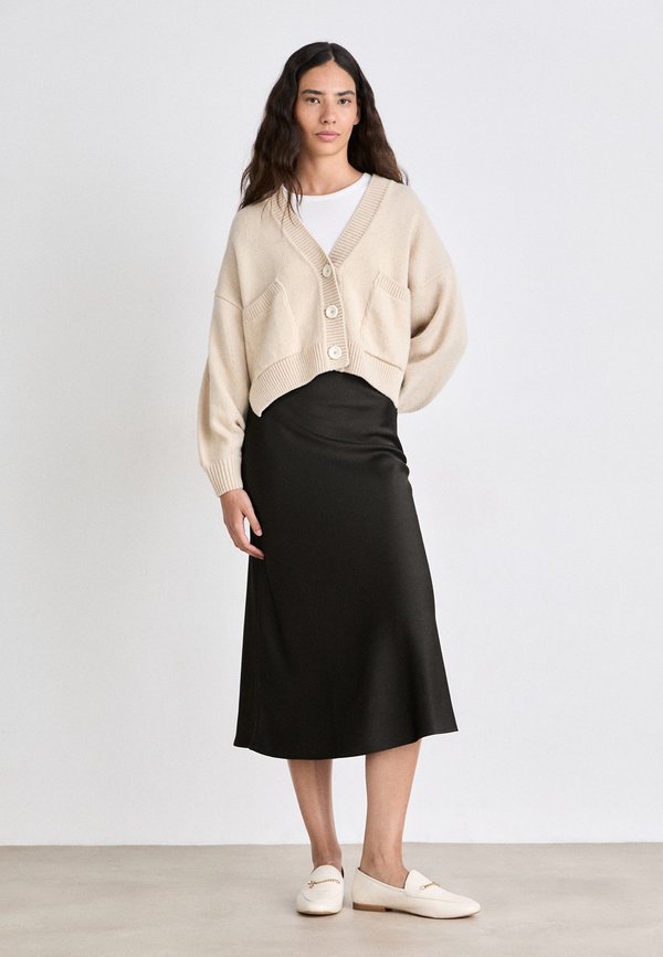 Vaalean beige lyhyt neuletakki, jossa on etutaskut ja ylisuuri istuvuus, päällä musta midi-hame. Valkoiset liukuvasti jalkaan menevät kengät viimeistelevät asun.