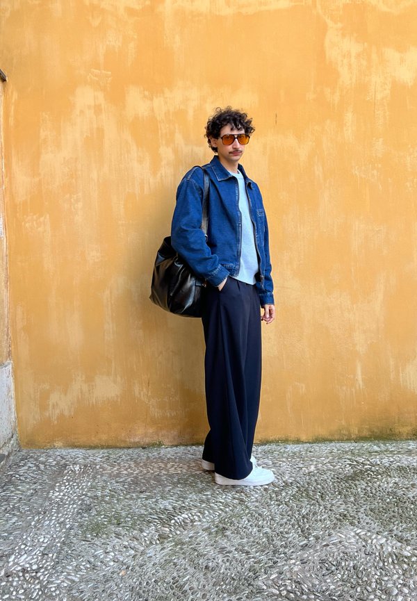 Chaqueta de mezclilla, suéter azul claro, pantalones de pierna ancha azul marino, bolso negro y zapatillas blancas. Cabello rizado oscuro y gafas de sol. Pared texturizada de fondo.