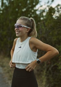 Idrettslig crop tank topp i lysegrå med mesh tekstur og logo. Sorte høytlivs leggings, klare solbriller med rosa tint, og et armbåndsur.