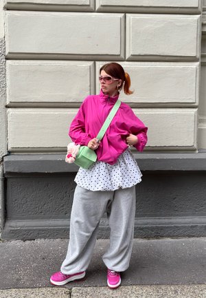 Rožinė užtrauktuku užsegama striukė, balta palaidinė su taškeliais, pilkos plačios kelnės, rožiniai sportbačiai, žalia per petį nešama rankinė su pritvirtintu žaislu.