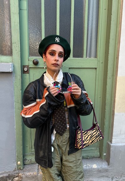 Chaqueta de cuero negra con rayas naranjas, boina verde, corbata estampada, pantalones verdes holgados, sosteniendo gafas de sol coloridas, bolso con estampado de cebra.