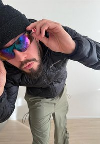 Homme barbu portant un bonnet noir et une veste ajustant des lunettes de soleil réfléchissantes colorées à l'intérieur, avec des murs et un sol de couleur claire visibles.