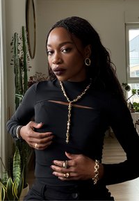 Haut noir ajusté à manches longues avec découpe, collier doré à perles, boucles d'oreilles créoles dorées et plusieurs bagues. Intérieur avec plantes vertes.