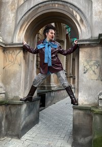 Osoba nosi vínovočervený kožený kabát, modrý šál, nohavice s zebrovým vzorom a hnedé čižmy, stojí rozkročená na dvoch kamenných rímsach pod oblúkom.