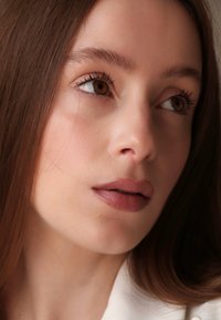 Nærbilde av et ansikt med glatt hud, langt brunt hår og nøytral sminke. Kjennetegn inkluderer brune øyne, definerte øyenbryn og myke rosa lepper.