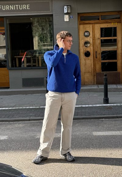 Suéter azul acanalado con cremallera de un cuarto, pantalones beige y zapatos deportivos negros. De pie en una calle con una puerta de madera y una tienda de muebles detrás.