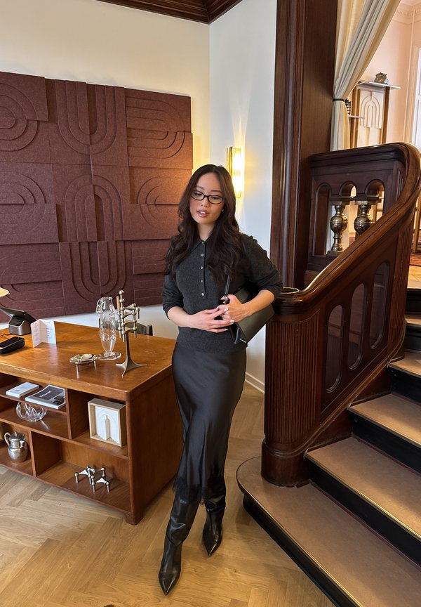 Femme aux longs cheveux foncés et lunettes, portant un haut et une jupe sombres, se tient près d'un escalier en bois et d'un buffet de style milieu du siècle dans un intérieur chaleureux.