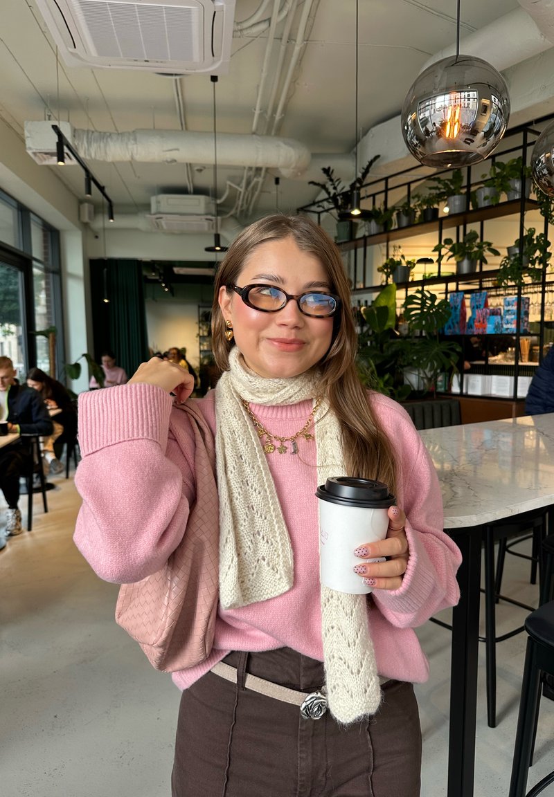 Maglione oversize rosa chiaro, sciarpa lavorata a maglia avorio, pantaloni marroni, borsa rosa, in mano una tazza di caffè usa e getta, unghie decorate, interno moderno.