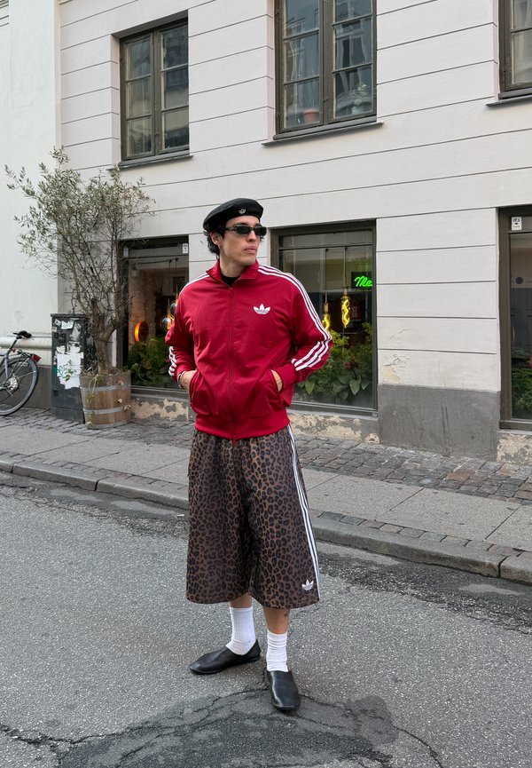 Jeune personne en veste Adidas rouge, shorts larges imprimés léopard, béret noir, lunettes de soleil, chaussettes blanches et chaussures noires debout dans une rue de la ville.