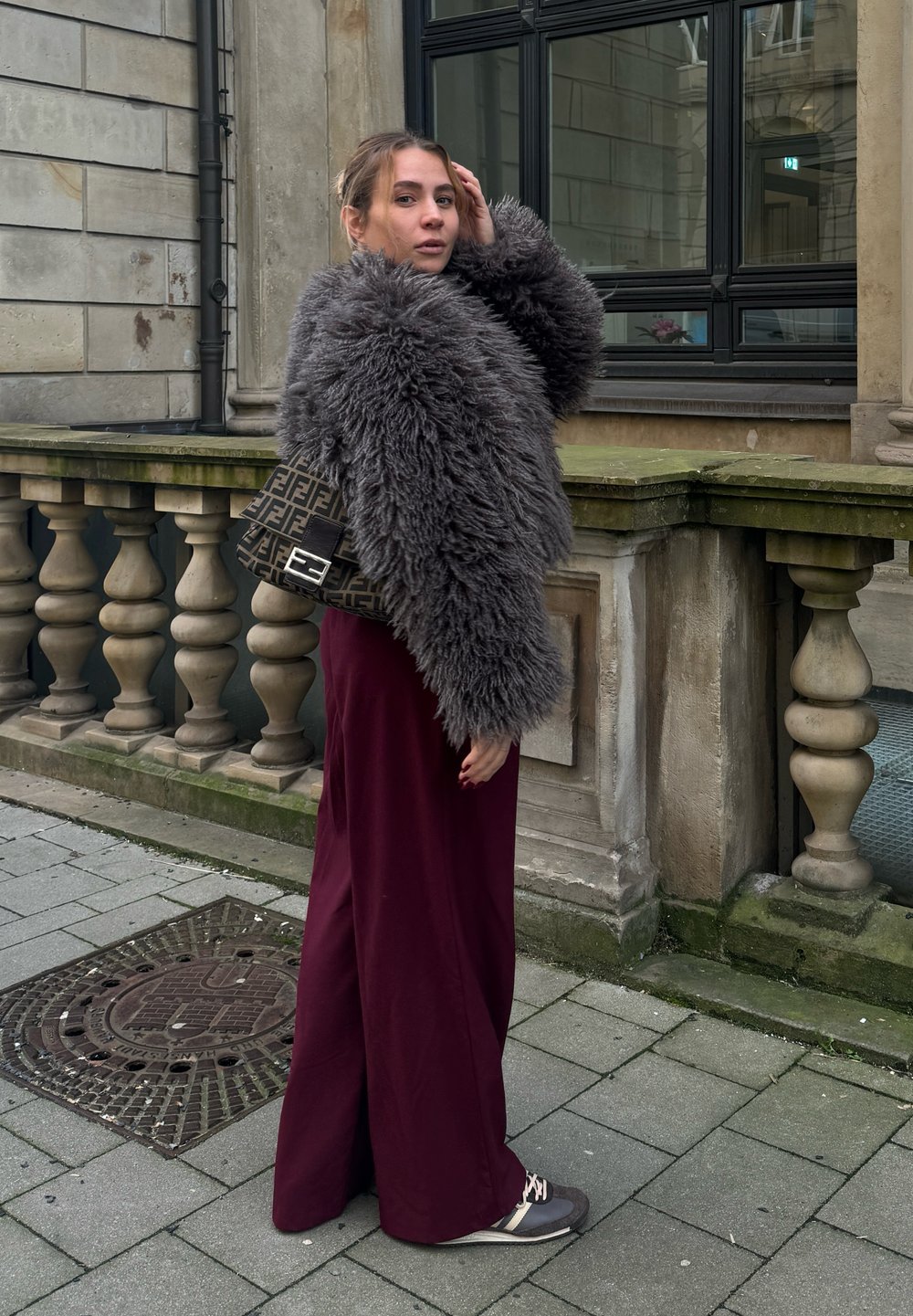 Γκρι ψεύτικο γούνινο μπουφάν, μπορντό παντελόνι σε φαρδιά γραμμή και μια τσάντα με σχέδιο μαύρου χρώματος. Το σκηνικό περιλαμβάνει πέτρινη αρχιτεκτονική και πλακόστρωτο.