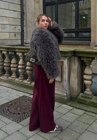 Γκρι ψεύτικο γούνινο μπουφάν, μπορντό παντελόνι σε φαρδιά γραμμή και μια τσάντα με σχέδιο μαύρου χρώματος. Το σκηνικό περιλαμβάνει πέτρινη αρχιτεκτονική και πλακόστρωτο.