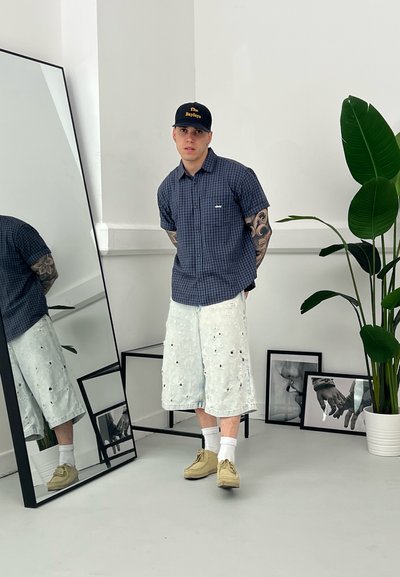 Camisa de cuadros azules de manga corta, pantalones cortos de mezclilla claros con detalles de flores, zapatos beige y calcetines blancos. De pie frente a un espejo.