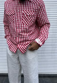 Camisa de botão às riscas vermelhas e brancas, com dois bolsos no peito e detalhes decorativos nas mangas, combinada com calças em tom claro e texturizadas.
