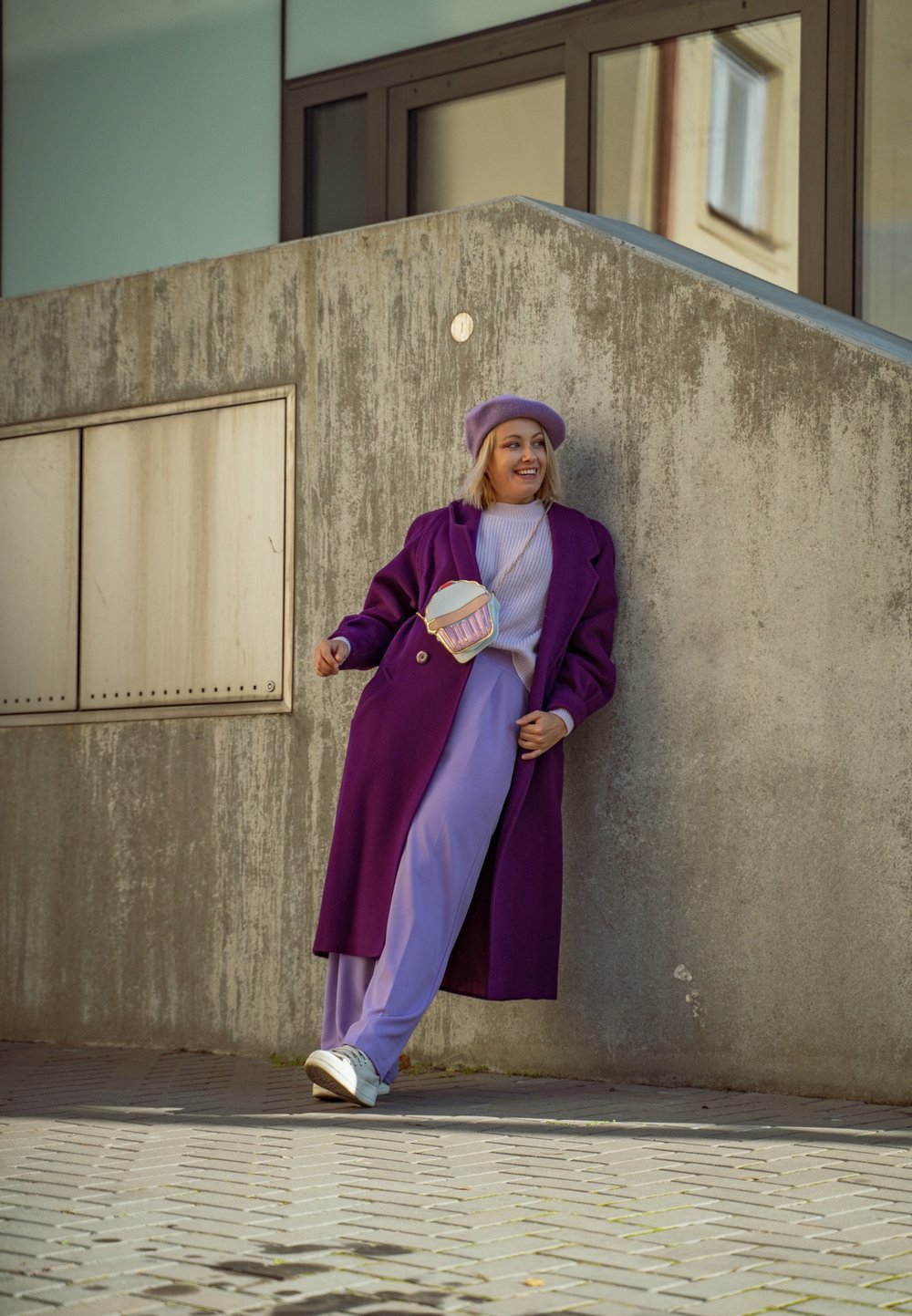 Μωβ παλτό, λεβάντα ρούχο και μπερέ. Λευκά αθλητικά παπούτσια και τσάντα σε σχήμα αλλάκας παστέλ. Υφή από σκυρόδεμα στο παρασκήνιο.