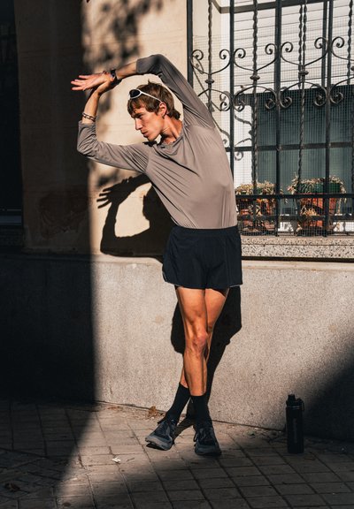 Hombre con ropa deportiva estirando los brazos por encima de la cabeza, de pie en una acera soleada cerca de un edificio con ventana con rejas y botella de agua en el suelo.