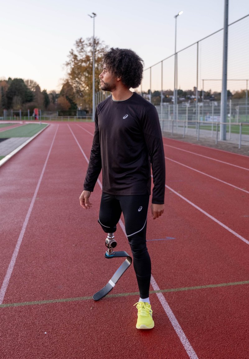 Atleta vestindo uma camisa preta de manga comprida e leggings pretas, com uma perna protética de fibra de carbono e sapatos desportivos amarelos brilhantes, de pé numa pista.