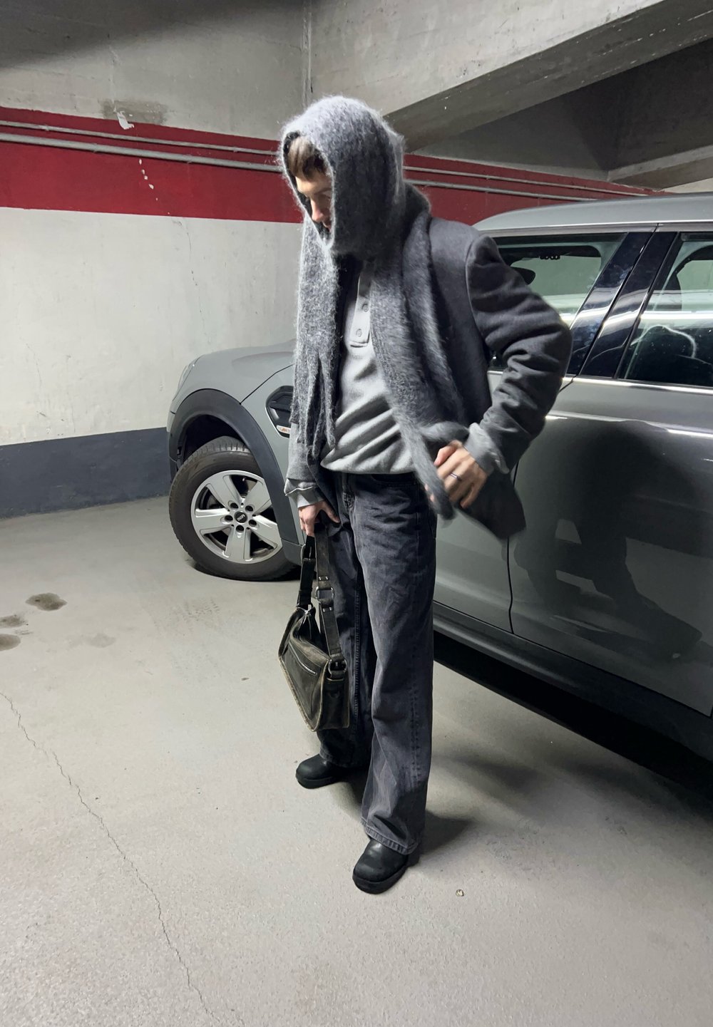 Suéter cinza oversized com um capuz texturizado e peludo, calças pretas e uma bolsa de ombro preta, de pé ao lado de um veículo cinza numa garagem de estacionamento.