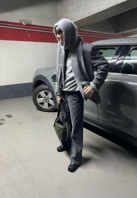 Suéter cinza oversized com um capuz texturizado e peludo, calças pretas e uma bolsa de ombro preta, de pé ao lado de um veículo cinza numa garagem de estacionamento.