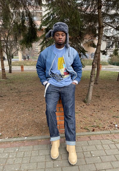 Chaqueta bomber de mezclilla en azul claro, gorro de punto gris, camiseta gráfica con un estampado amarillo, jeans azul oscuro y botines beige con cordones.