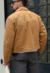 Barna velúr dzsekis férfi, hátán "Queens" hímzés, fehér táska pántot tart a kezében, egy fekete kovácsoltvas kapu előtt áll.