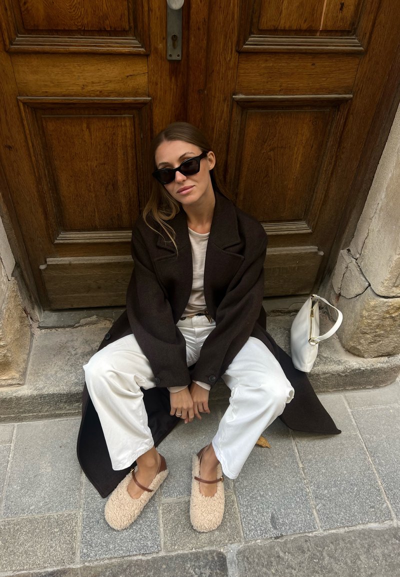Dark brown wool coat, white t-shirt, white wide-leg trousers, and tan fluffy slippers with brown straps. A stylish white handbag is included in the frame.