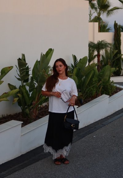 Camiseta blanca, falda negra hasta los tobillos con borde de encaje floral, bolso negro con herrajes dorados, de pie cerca de plantas verdes en un sendero.