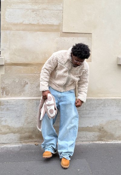 Hombre con chaqueta beige de peluche, jeans holgados azul claro y botas marrones, de pie contra una pared de piedra, sosteniendo una manta doblada con estampado.