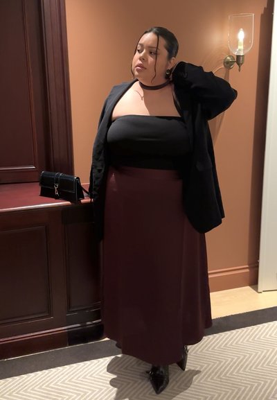 Mujer de pie en el interior, vistiendo un top negro sin tirantes, blazer negro, falda burdeos y botas negras puntiagudas, con un bolso negro sobre un mostrador de madera.