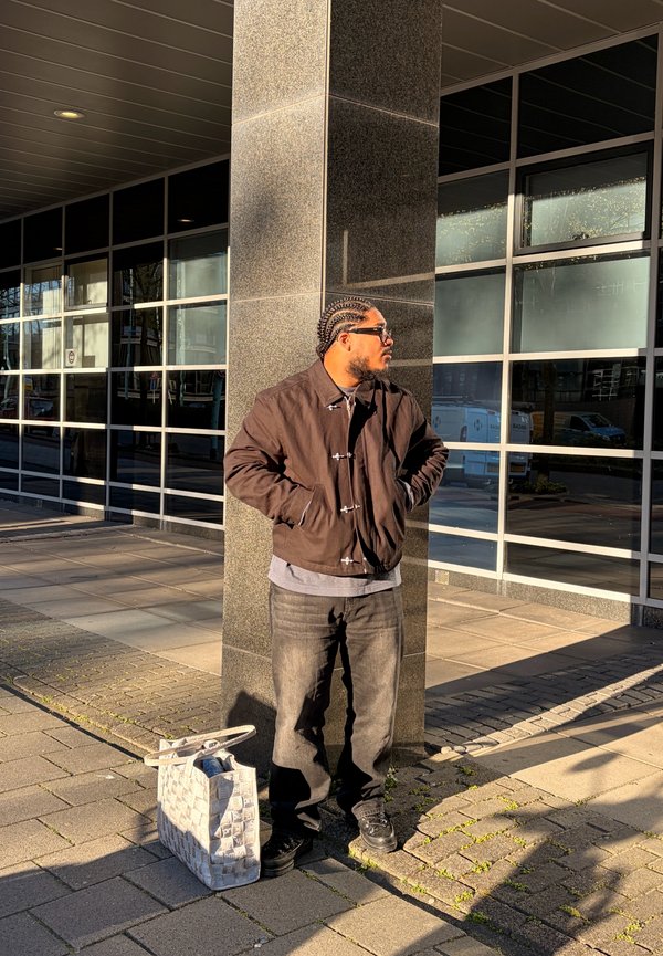 Man with braided hair and sunglasses stands with hands in pockets by a building pillar, next to a patterned tote bag on the pavement.